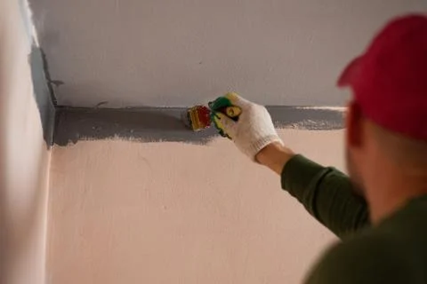 A man, a construction worker in a red cap, paints the wall with a brush with Stock Photos