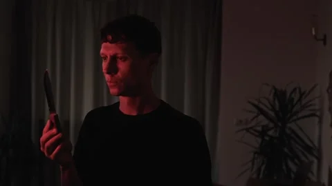 Man contemplating revenge while examining a knife at home, reflecting on a 스톡 동영상 347300625