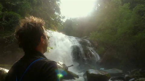 Man contemplating serene waterfall in lush forest under bright Stock Footage 280176982