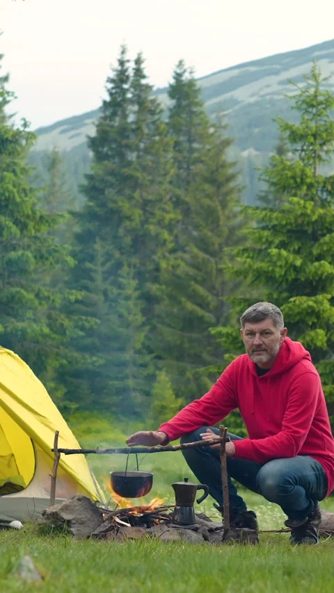 Man cooking food over campfire near tent during outdoor camping in mountains Stock Footage 336431453