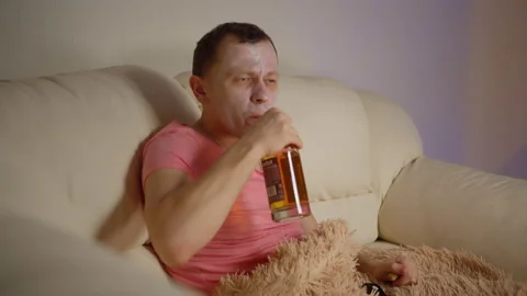 Man in a cosmetic mask sits on the sofa watching TV, opens a bottle of alcohol Stock Footage 137901331