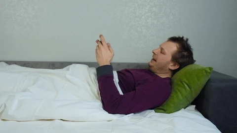 Man covered with a blanket is lying on the bed and playing a mobile game Stock-Footage 129363949