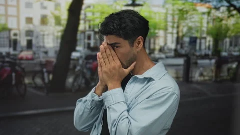Man covers face with both hands in denim jacket on crowded urban street bes.. 스톡 동영상 313864970