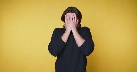Man covers his face with his hands in a simple yellow background while expr.. Video stock 328249952
