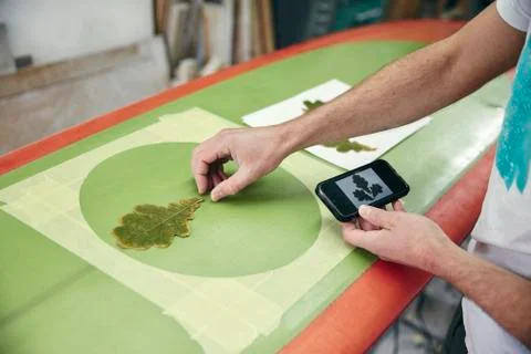 Man creating a design on surfboard laying an oak leaf on the board, using mobile Stock Photos