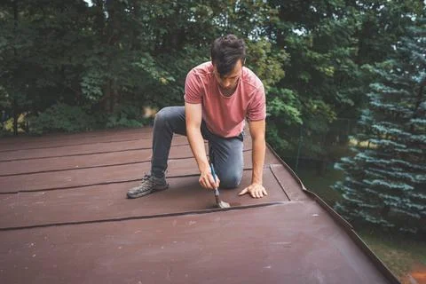 Man crouches down, carefully painting the surface of a roof with a small brus Stock Photos