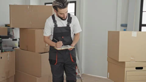 Man crouching and writing on a clipboard beside stacked cardboard boxes in .. Stock Footage 337371701