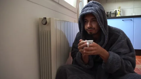 A man with a cup of hot drink in a warm bathrobe sits by a cold radiator. No gas Video stock 221028390