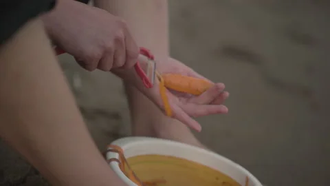 A man cuts the peel from a carrot while sitting on the sand Stock Footage 142189229