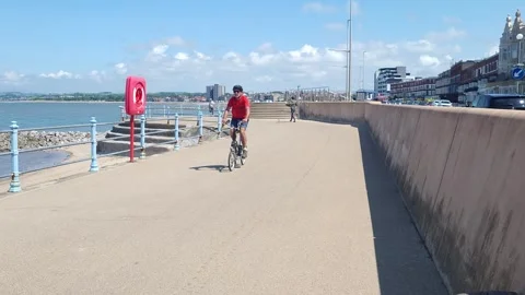 Man cycles towards camera on Brompton folding bike, Morecambe seafront, UK Stock Footage 244325328