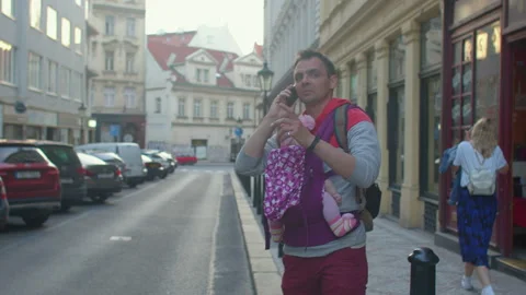 Man dad holding small child in sling. He Uses phone, makes a call to service. Video stock 156082183