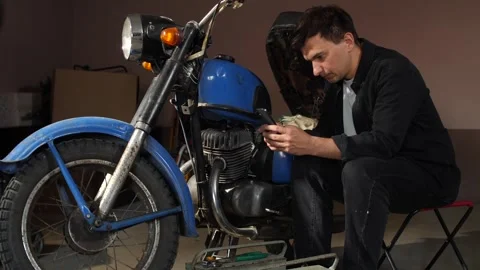 Man in dark clothes carefully inspecting a vintage blue motorcycle's engine Stock Footage 342769794