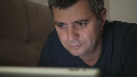 A man in a dark room lying on a couch smiling looking at an electronic tablet Stock Footage 92925247