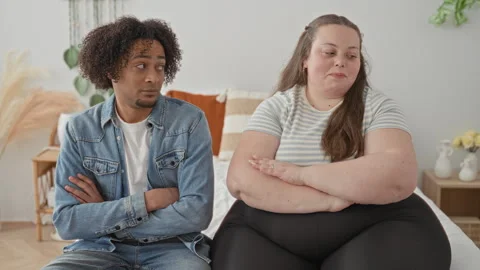 Man in denim jacket and plus size woman sit side by side on a bed, arms cro.. Stock Footage 337389463
