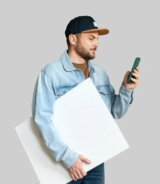 Man in denim jacket and snapback holding blank white board while using smartp Stock Photos