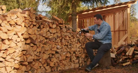 Man in a denim jacket sitting and holding a thermos by a large woodpile. Rural Stock Footage 265007808