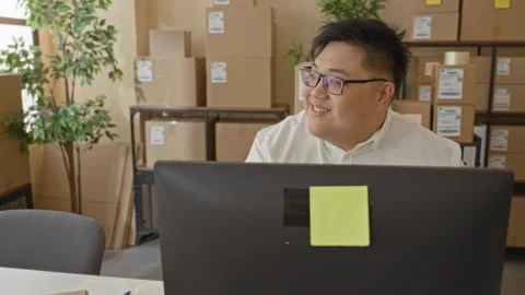 Man at desktop computer smiles while looking to right among stacked parcels.. Stock Footage 347839805