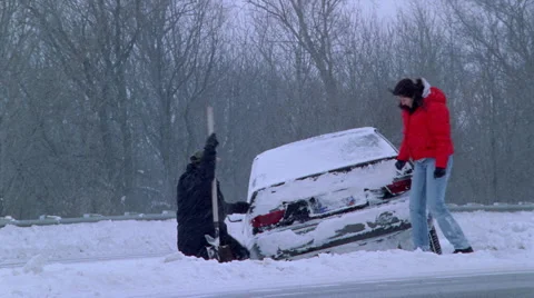 Man digging out a car stuck in snow beside a highway while woman looks on Vídeos de archivo 59262734