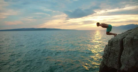 Man diving off rocky cliff into ocean at sunset with scenic horizon and calm Stock Footage 320090213
