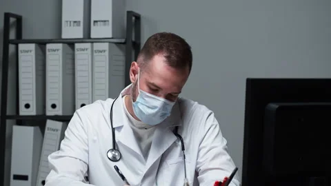 Man doctor wearing protective face mask, medical gown sitting and writing Stock Footage 158619683