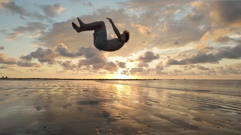 Man does a back somersault backflip in front of a beach sunset Stock Footage 302691309
