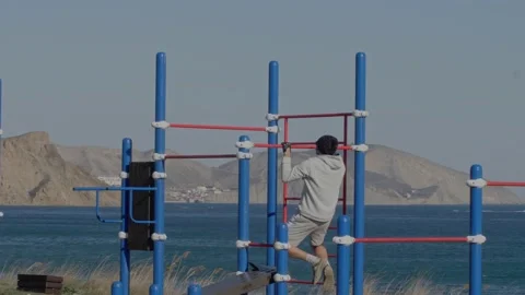 Man does gymnastic exercises on a turnstile by the sea on a sunny day Stock Footage 131090253