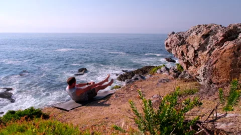 Man does seated abdominal plank hold core flutter kicks on sea cliff in morning Stock Footage 322887742