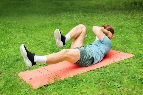 A man is doing bicycle crunches on an orange mat in a green park. This outd.. Stock Photos