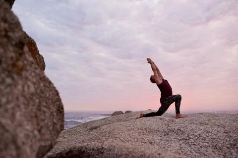 Man doing the cresent lunge pose on rocks at dusk Stock Photos