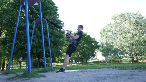 Man doing intense pull-up exercising on fitness loops in windy summer park. 4K Stock Footage 99494658