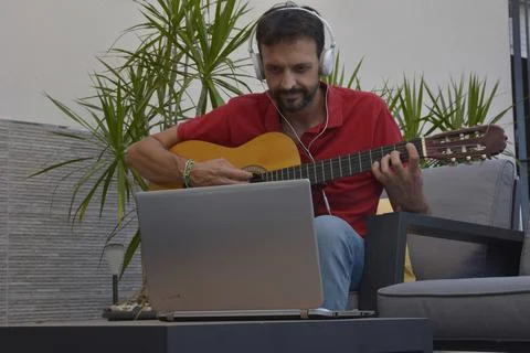 Man doing an online course to learn to play classical guitar. He is sitting o Stock Photos
