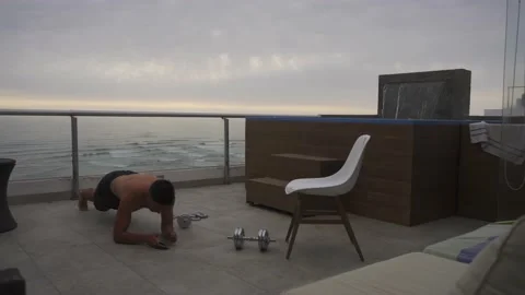 A man doing planks and timing with his watch on a terrace with dumbbells on the Stock Footage 186213788
