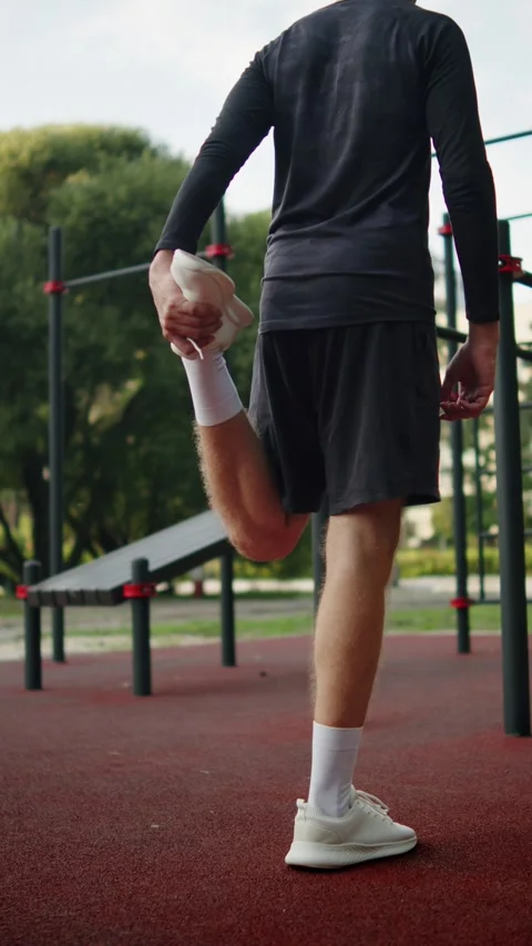 Man doing a standing quadriceps stretch on an outside gym. Man in black ath.. Video stock 341728209