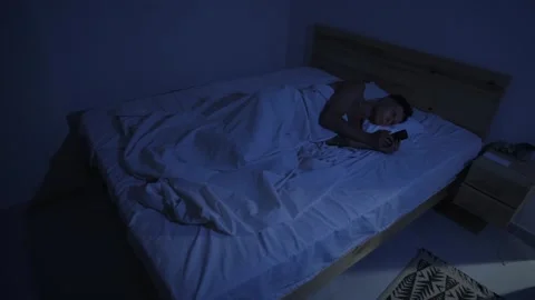 Man doomscrolling on smartphone in bed late at night, delaying sleep due to Stock-Footage 346248410