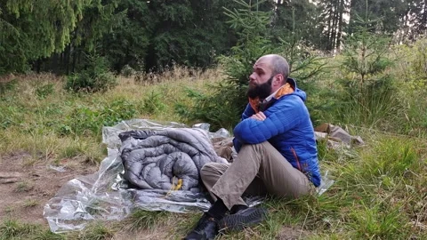 Man in down jacket sitting near sleeping bag in pine forest looking up Stock Footage 344893588