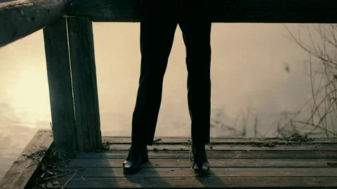 A man dressed in a black elegant suit stands on a wooden bridge at sunset. Stock Footage 245486049