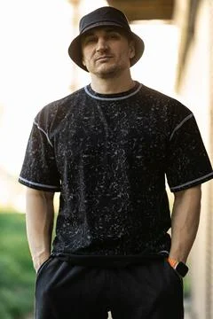Man dressed in a black patterned shirt and bucket hat stands with his hands in Stock Photos