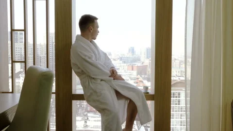 Man dressed in white bathrobe drinking coffee, looking out of window at home. 4K 動画素材 107935376
