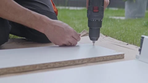 Man drills hole in drawer panel on cardboard with grass in the background Video stock 335661560