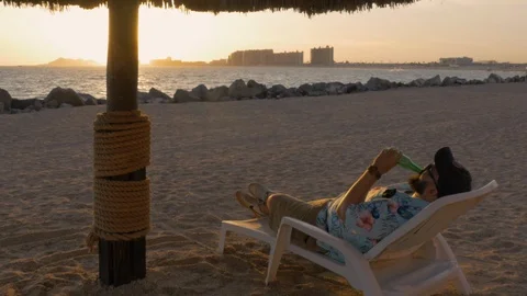 Man Drinking a Beer on the Beach at Sunset Under a Palm Umbrella Stock Footage 101265909