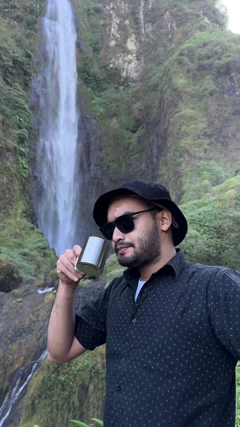 Man Drinking Beverage from Metal Mug by Waterfall in Mountains Vertical Video stock 323913414