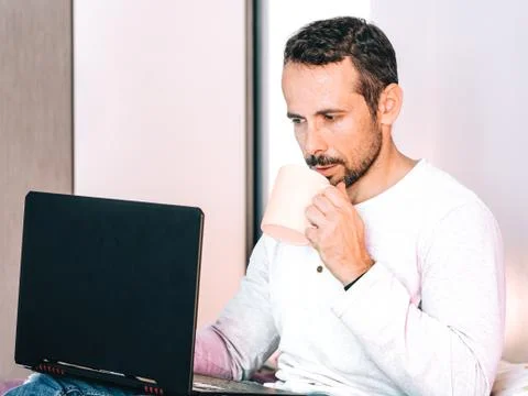Man drinking a cup of coffee and working on his computer in the bedroom. Stock Photos