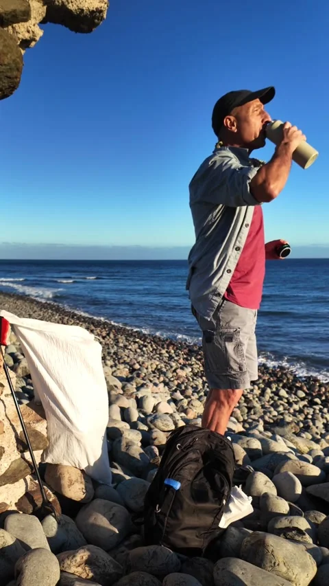 Man drinking fresh water from his thermal bottle on a rocky beach Vídeos de archivo 329242364