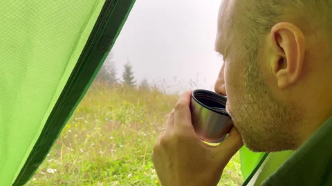 Man drinking hot tea inside camping tent and enjoying autumn rainy morning in mo Stock Footage 300578980