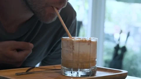 Man drinking iced creamy coffee with straw on wooden tray in cozy cafe settings Stock Footage 312813986