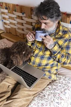 Man drinking tea and using laptop with dog in bed Stock Photos