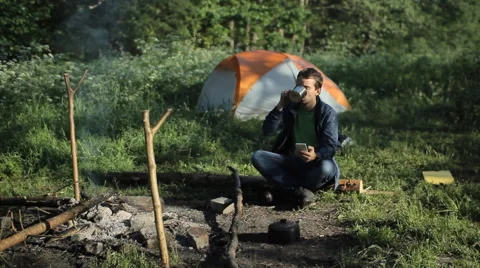 The man is drinking tea near the fire and taking pictures the forest and tent Stock Footage 64310077