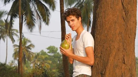 Man drinks a coconut through a straw while standing by a palm tree 写真素材