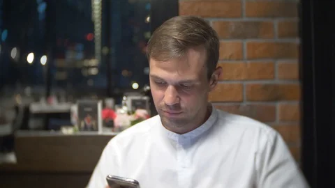 A man drinks coffee, checking a cell phone at night in a restaurant. he looks at Видео 86440181