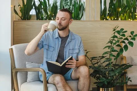 Man drinks coffee while reading book in modern workspace with plants during.. Stock Photos